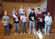 Bērni stāv uz skatuves kāpnēm, rokās diplomi