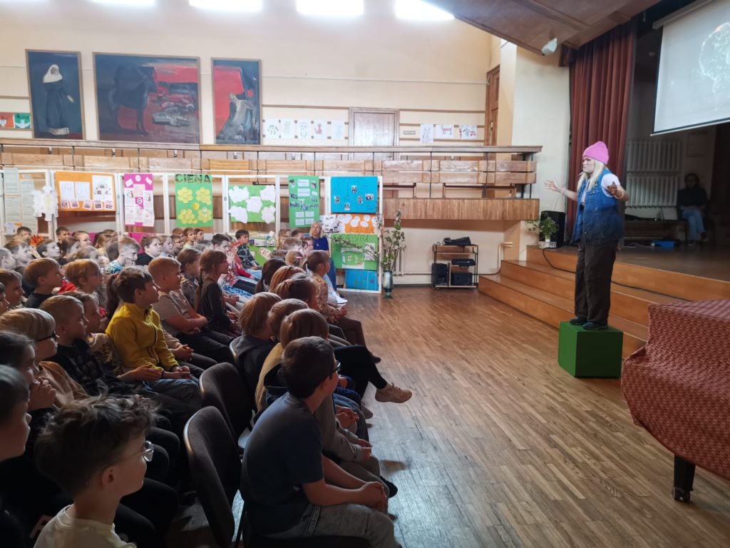 Meitene (aktrise) stāv uz zaļas koka kastes, bērni sēž un skatās, aktu zāle, skola