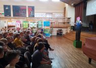 Meitene (aktrise) stāv uz zaļas koka kastes, bērni sēž un skatās, aktu zāle, skola
