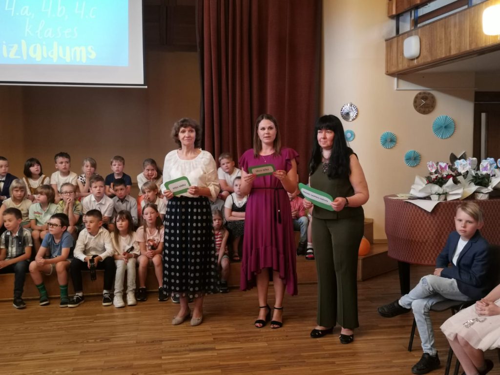 Aktu zāle, trīs skolotājas, bērni sēž uz podestiem.