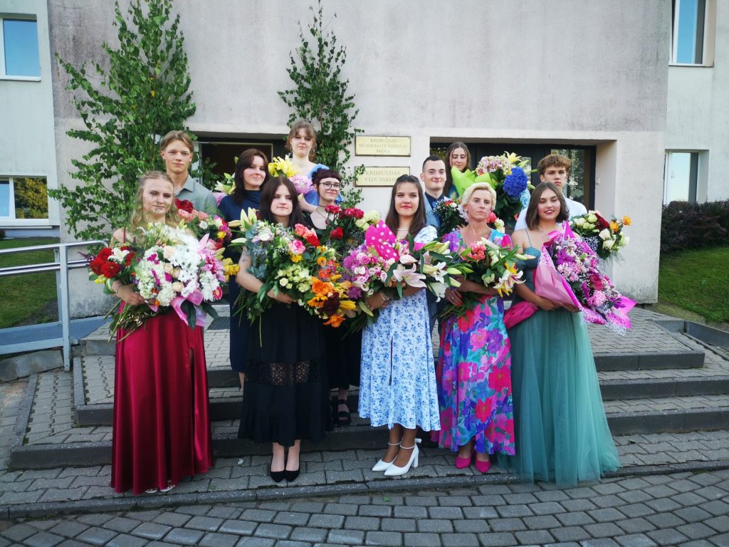 Skolas priekša, cilvēki sapucējušies ar ziediem stā, smaida, 12.klases izlaiduma klase
