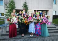 Skolas priekša, cilvēki sapucējušies ar ziediem stā, smaida, 12.klases izlaiduma klase