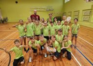 Skolēni un skolotājs sporta zālē, zaļos krekliņos priecīgi pozē