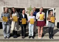 Skolēni ar zelta liecībā, stāv rindā skolas priekšā
