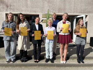 Skolēni ar zelta liecībā, stāv rindā skolas priekšā